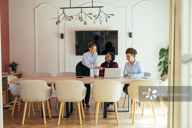 三位不同背景的女商人在现代会议室的会议桌上合作，旁边有一台笔记本电脑。图片素材
