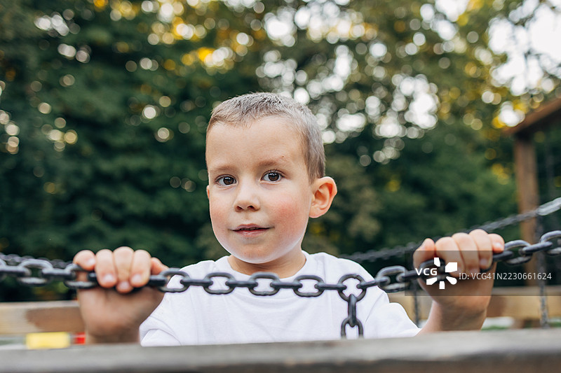 一个年轻男孩在游乐场玩耍，抓着链子并看着镜头。图片素材