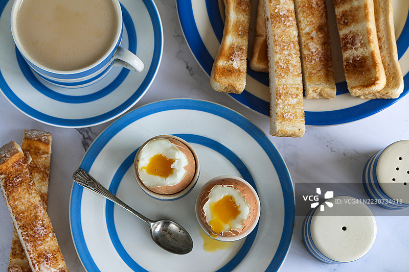 蓝白相间的康沃尔陶器，茶盘和盛有流心软煮蛋的蛋杯，白色涂黄油的烤面包士兵，刀子，盛有奶茶的茶杯和碟子，盐和胡椒瓶，俯视图图片素材