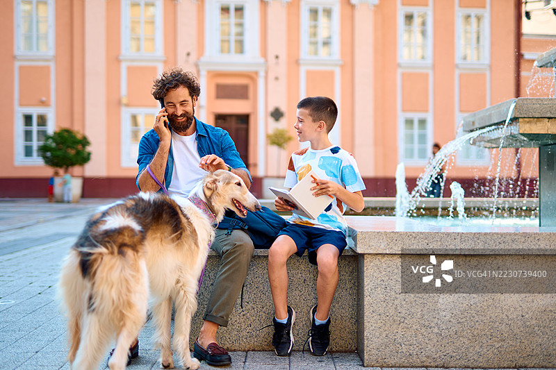 父亲和儿子在喷泉旁与他们的狗放松。图片素材
