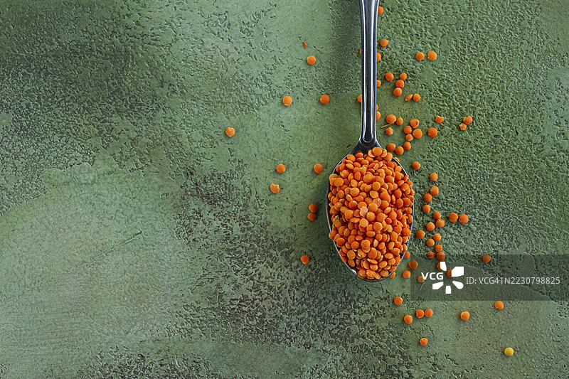 明亮的橙色扁豆从银色勺子中洒落在有纹理的绿色表面上，突显了这种多用途烹饪原料的鲜艳色彩和营养价值。图片素材