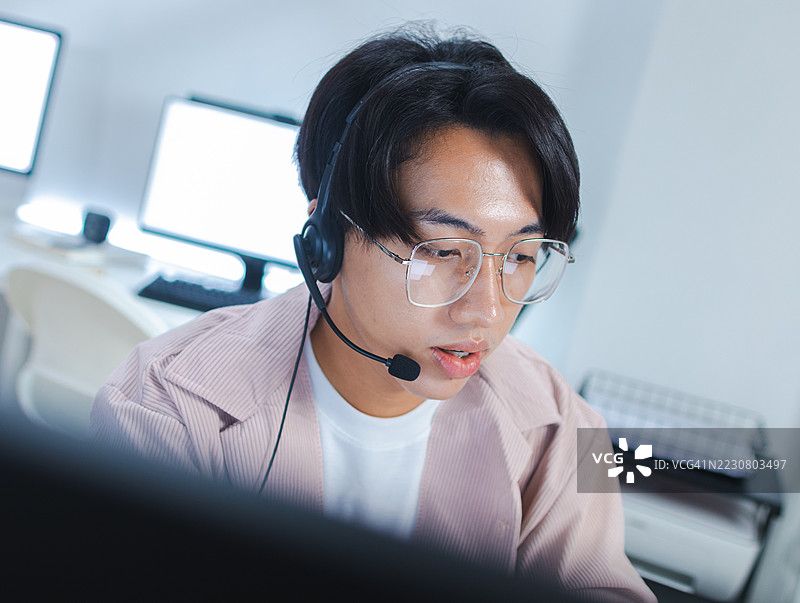 亚洲男性IT支持呼叫中心工作人员，佩戴耳机在办公室电脑前工作图片素材