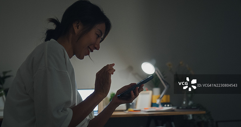 年轻的亚洲女性在夜晚的卧室里坐在床上，使用智能手机打开灯光。智能家居技术为日常生活提供便利。图片素材