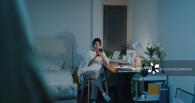 年轻的亚洲女性在夜晚的卧室里坐在床上，使用智能手机打开灯光。智能家居技术为日常生活提供便利。图片素材