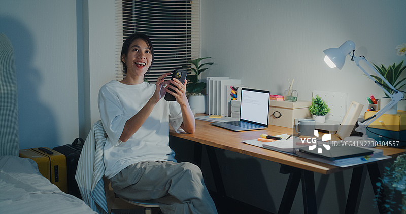 年轻的亚洲女性在夜晚的卧室里坐在床上，使用智能手机打开灯光。智能家居技术为日常生活提供便利。图片素材