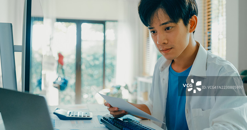 年轻亚洲男性在家工作，近距离观察他在电脑屏幕上分析财务数据，反映出现代家庭办公室的布局和远程工作的状态。远程工作与财务。图片素材