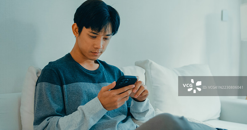 年轻的亚洲男子坐在沙发上，专注于使用智能手机，展现出一种随意而互联的家庭生活方式。远程工作与金融。图片素材