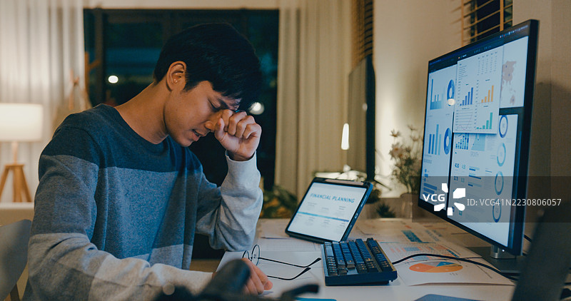 年轻的亚洲男性疲惫的员工在家中办公室加班，因财务规划和数据分析任务感到心理疲惫，面对多台屏幕感到不堪重负。远程工作与财务。图片素材