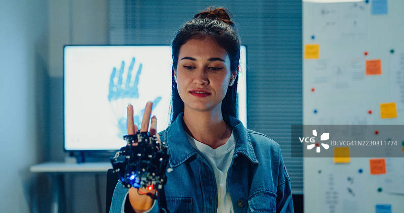 年轻女性工程师在夜晚的科技实验室里加班，微笑着看向镜头，测试机器人手臂原型。机器学习、创新与创造力。人工智能与机器人开发。图片素材