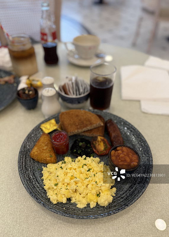 早餐餐桌上摆放着炒鸡蛋、烤豆、全麦吐司、土豆饼、蘑菇和番茄，配有番茄酱，所有食物都在盘子里准备好享用，旁边还有一杯饮料，特写镜头。图片素材