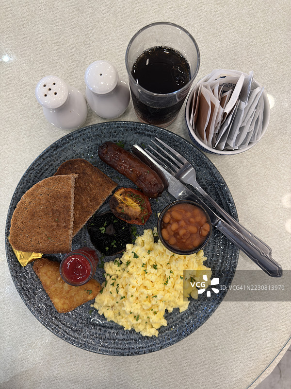 早餐盘上的炒鸡蛋、香肠、棕色吐司、土豆饼、蘑菇片和番茄，旁边还有一小碗烤豆和餐具，以及饮料的特写。图片素材