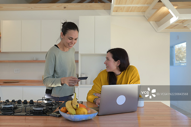 两位女士在厨房里使用平板电脑和笔记本电脑刷信用卡购物。图片素材
