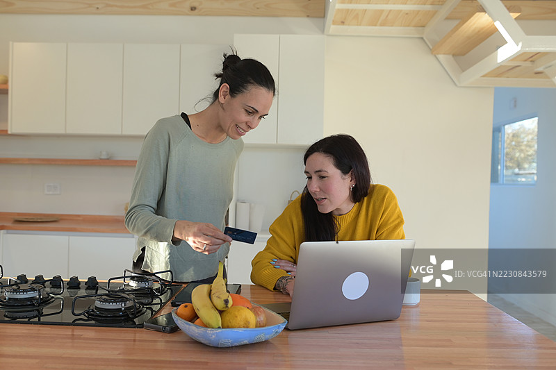 两位女性在厨房里使用平板电脑和笔记本电脑，并用信用卡购物。图片素材