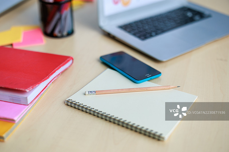 特写镜头展示了一个桌子，上面有一个螺旋笔记本、铅笔、多彩文件夹、智能手机和打开的笔记本电脑，代表学习、组织和现代工作空间的布置。图片素材
