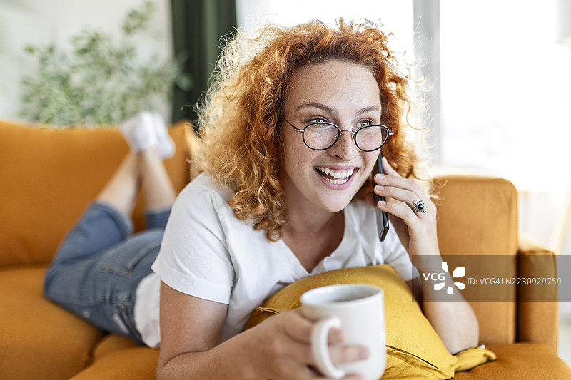 美丽的女人在舒适的沙发上放松，手里拿着咖啡和手机图片素材