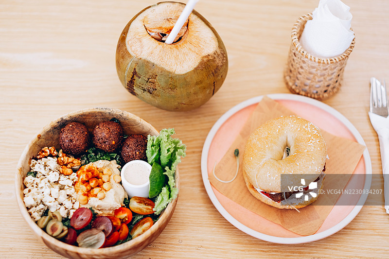 色彩缤纷的素食碗，里面有法拉费尔、鹰嘴豆泥、蔬菜和谷物，旁边是一份芝麻贝果三明治和一只带吸管的新鲜椰子。植物性餐饮、健康生活方式和素食烹饪理念。图片素材