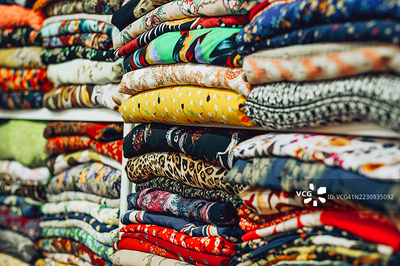整齐叠放在布料市场或纺织店中的五彩斑斓、图案各异的布料，展示了多样的质地、印花和文化风格。图片素材