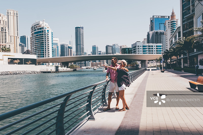 一对情侣在海滨长廊上漫步，背景是高耸的摩天大楼和一座桥。城市旅游、现代旅行和爱情的概念。图片素材