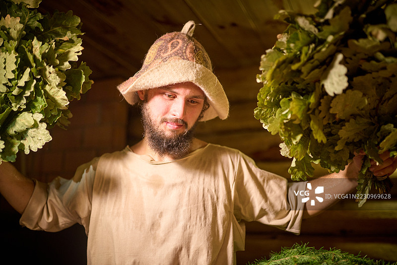 年轻的男浴室服务员手持两把橡木扫帚图片素材