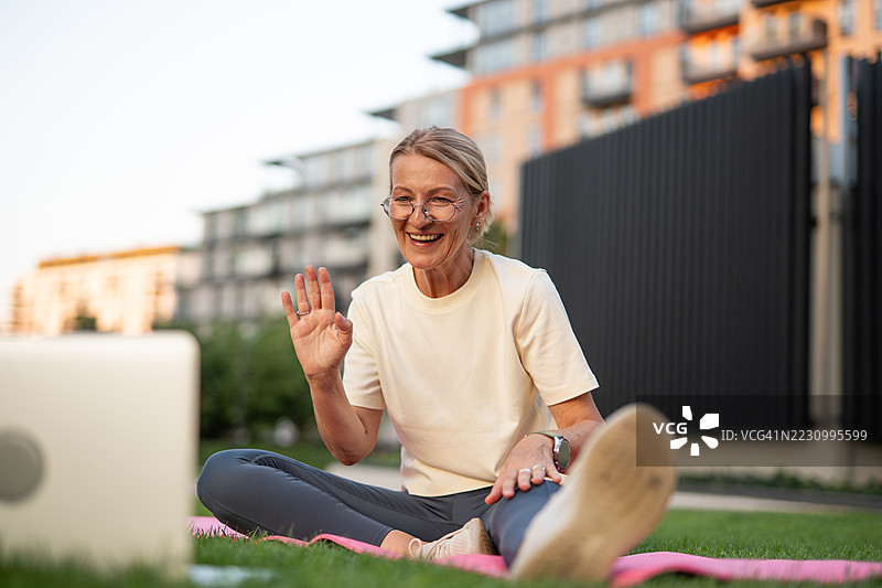 户外在线瑜伽课上，年长女性对着笔记本电脑挥手。图片素材