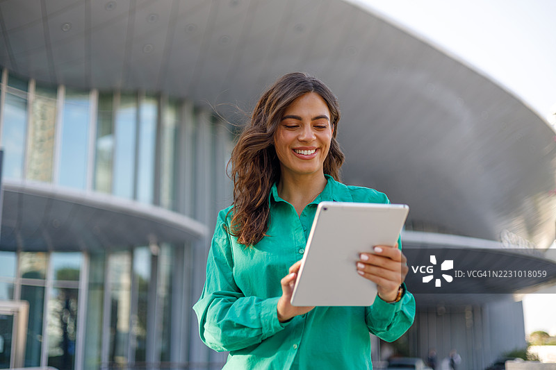 快乐的女商人微笑着使用平板电脑，自信地站在一座现代化办公大楼前，天空晴朗。图片素材