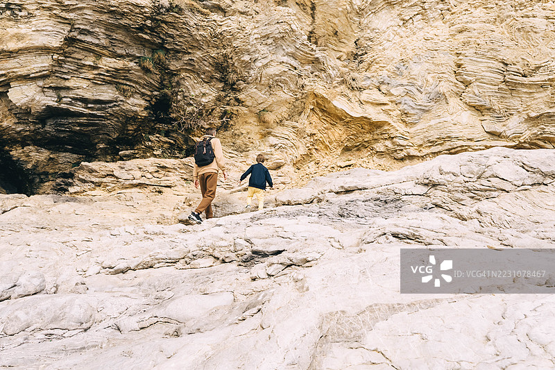 年轻父亲背着背包，带着小男孩在岩石海滩上散步。图片素材
