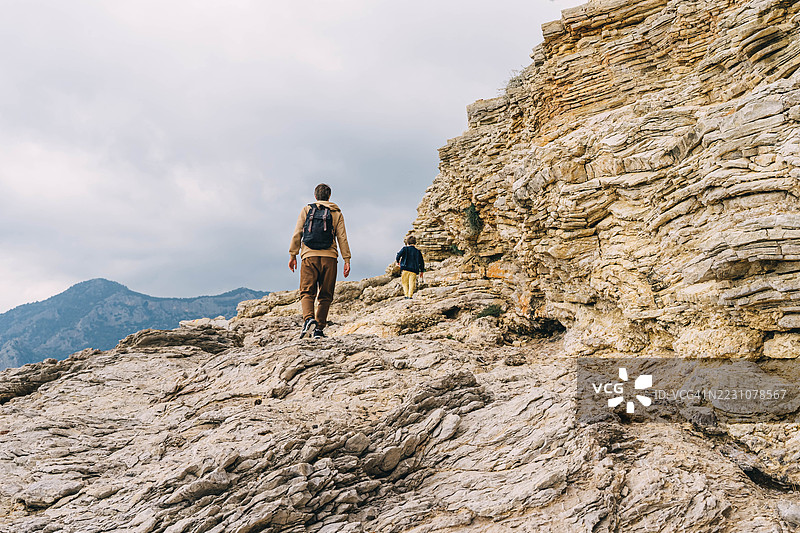 年轻父亲背着背包，带着小男孩在岩石海滩上散步。图片素材