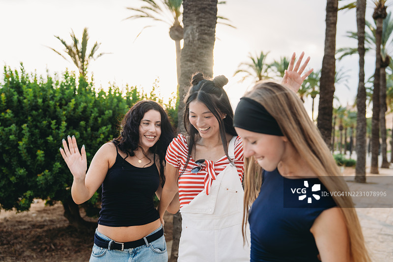 三位年轻女性在棕榈树环绕的公园中挥手微笑图片素材