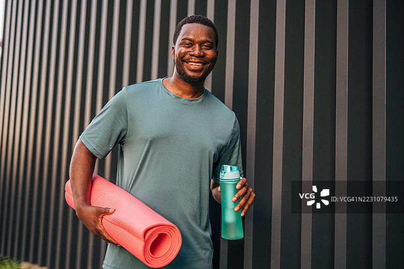 微笑的黑人男子在锻炼后手持瑜伽垫和水瓶图片素材