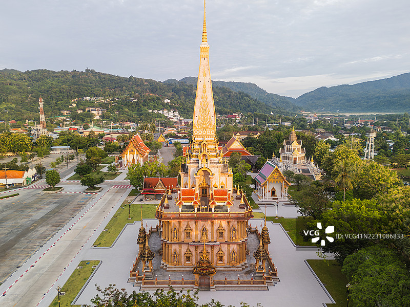 从空中俯瞰泰国普吉岛的查龙寺金色佛塔，周围环绕着郁郁葱葱的树木、寺庙和远处的山景。图片素材