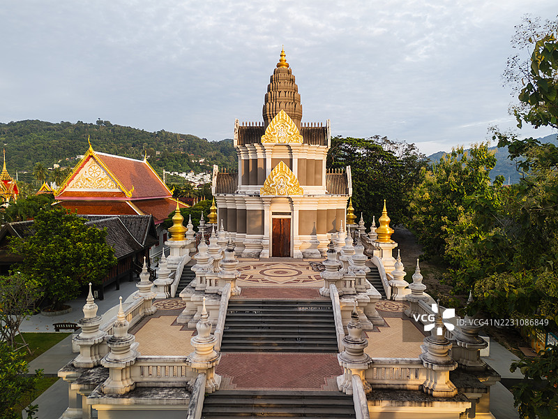 普吉岛一座精美设计的泰国寺庙的鸟瞰图，金色细节环绕着树木和山脉，天空多云。图片素材