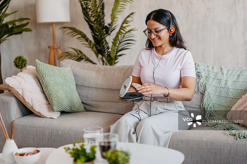 年轻的印度女性坐在沙发上，将CD放入便携式CD播放器中，身处客厅。图片素材