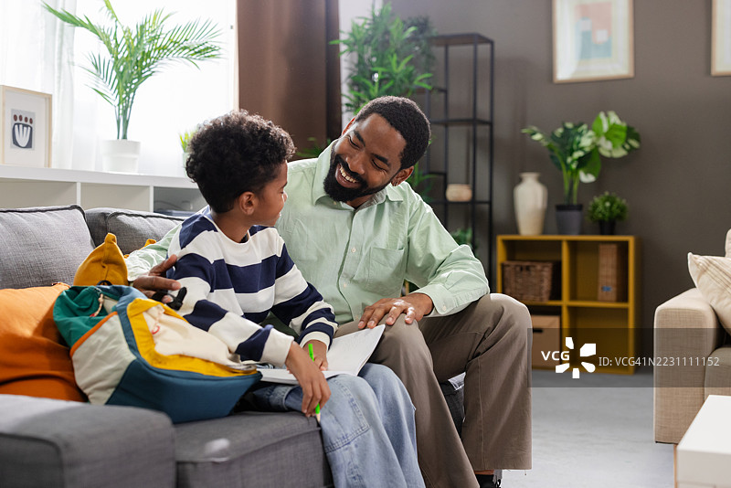 快乐的非裔美国父亲在家沙发上帮助儿子做作业图片素材