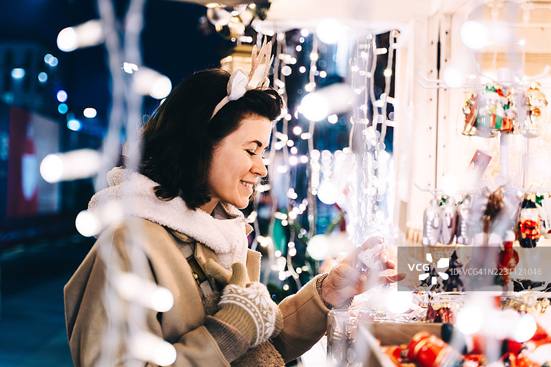 一位快乐的女性头戴驯鹿角，微笑着低头浏览圣诞市场摊位上的节日商品。图片素材