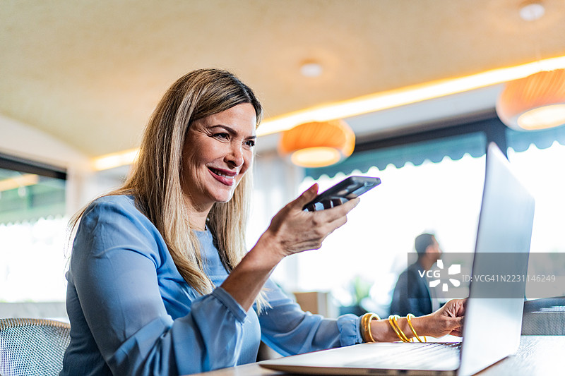 成熟的女商人在咖啡店使用笔记本电脑并通过手机发送语音信息图片素材