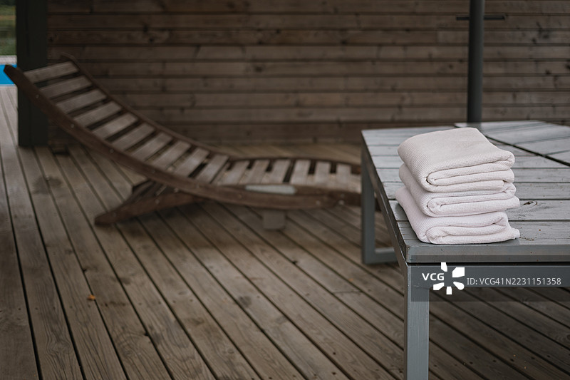 一堆干净的白色毛巾放在木制长椅上，旁边是一个木制日光浴椅，位于桑拿旁的露台上。图片素材
