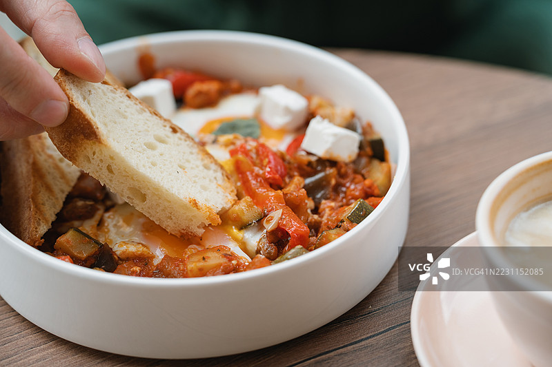 特写镜头：一名男子坐在桌旁，将一片面包浸入装有鸡蛋的沙克舒卡盘中。图片素材