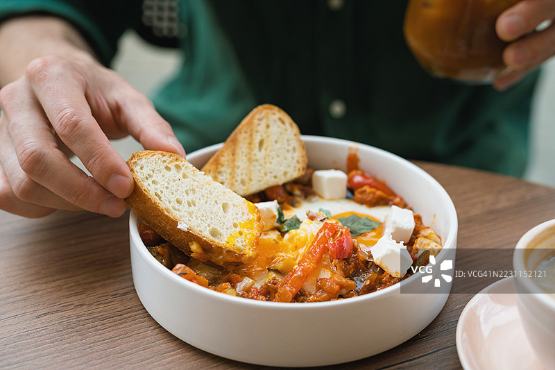 特写镜头：一名男子坐在桌旁，将一片面包浸入装有鸡蛋的沙克舒卡盘中。图片素材