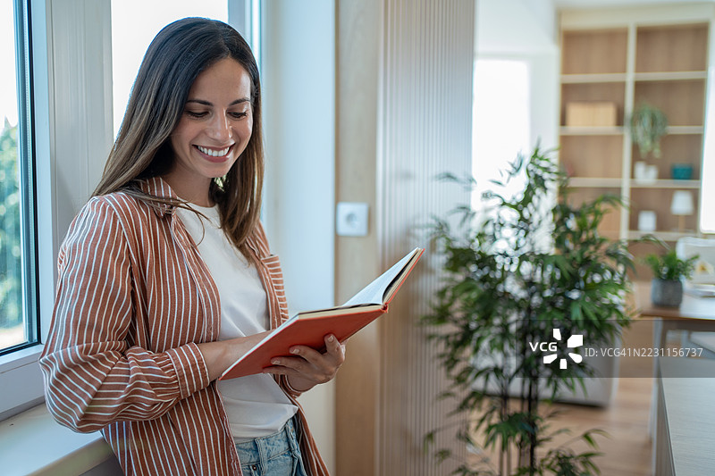 年轻女性在窗边阅读一本书并微笑图片素材