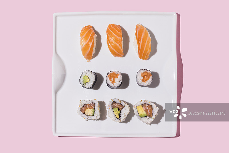 白盘上的寿司握饭和卷寿司，配有米饭、鳄梨、生鱼和三文鱼，背景为粉色。日本、亚洲、健康、富含蛋白质和碳水化合物的食物。图片素材