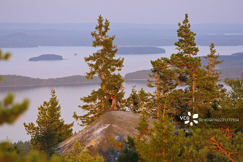 一个人坐在岩石上，周围是松树，俯瞰着日落时分宁静的湖泊。图片素材