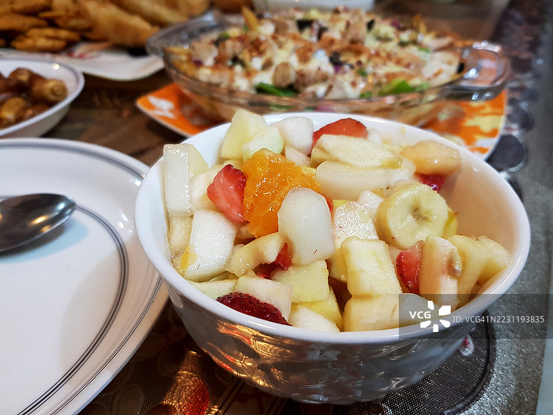 餐桌上的碗里盛着混合水果沙拉，背景中还有其他食物。图片素材