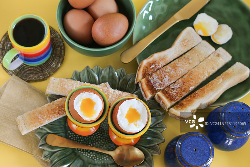 早餐场景的图片，绿色向日葵盘，两个蛋杯，流心的软煮蛋，涂了黄油的白色吐司士兵，金属茶匙和刀，蓝色盐和胡椒罐，一碗鸡蛋，彩虹条纹的浓缩咖啡杯，黄色背景，俯视角度。图片素材