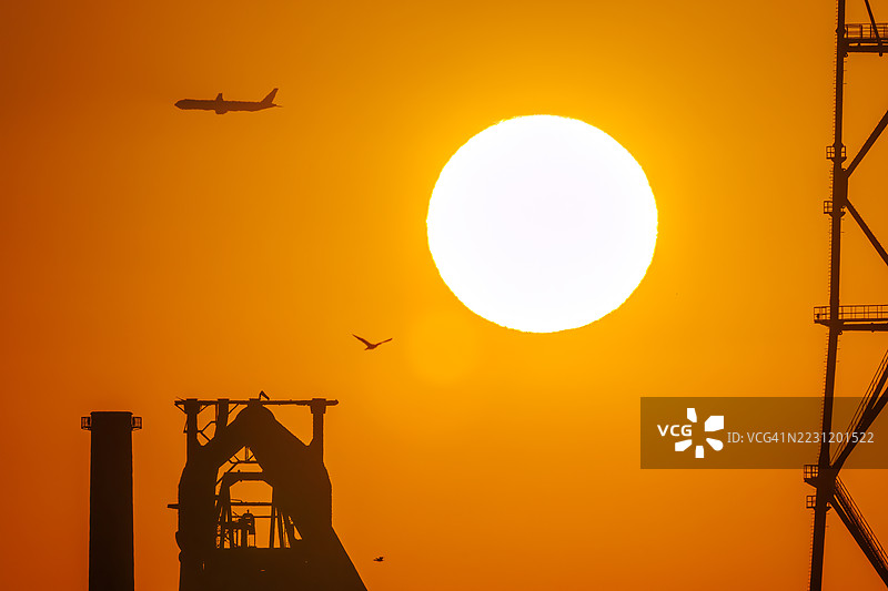 第一缕阳光与一架客机和鸟的剪影。它即将降落在附近的羽田机场。第一缕阳光从运河对岸的烟囱和电厂、钢铁厂上升。横滨，神奈川。图片素材