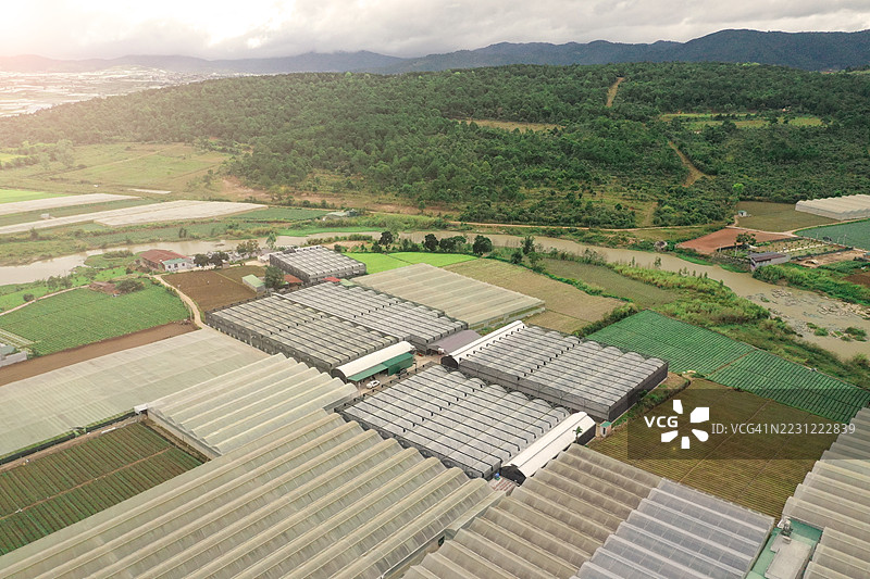 越南农村地区大规模温室和农田的鸟瞰图。农业温室的排成行，周围环绕着田野、河流和森林山丘，展示了东南亚现代农业和园艺的景观。图片素材