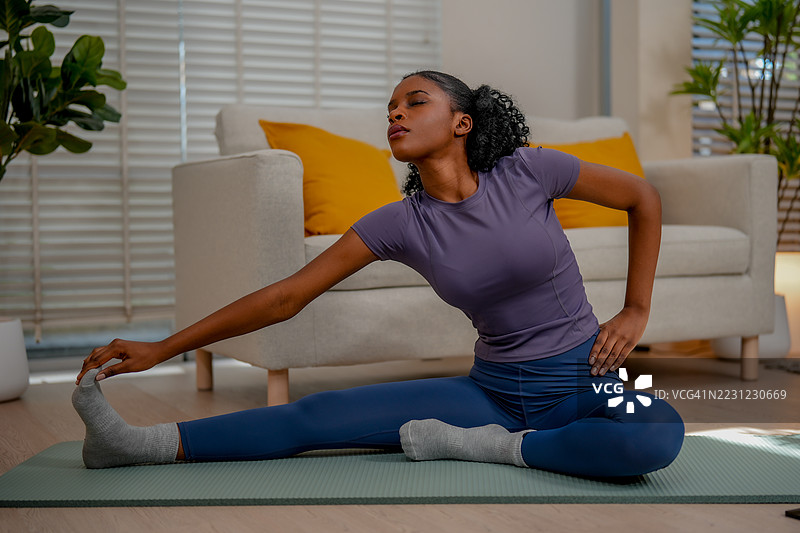 黑人女性在室内锻炼，专注地练习瑜伽，伸展腿部，坐在垫子上。这种宁静的氛围促进了健康和正念。图片素材