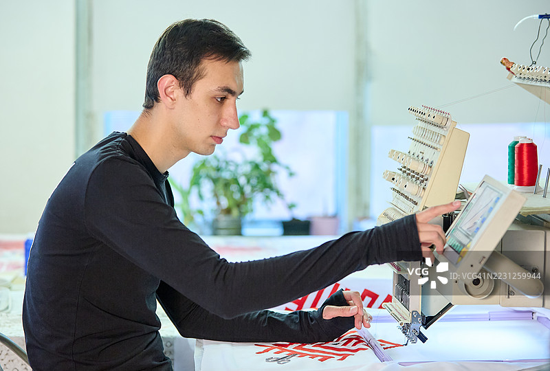 年轻男设计师正在设置一台电脑刺绣机，将传统图案刺绣到白色布料上。图片素材