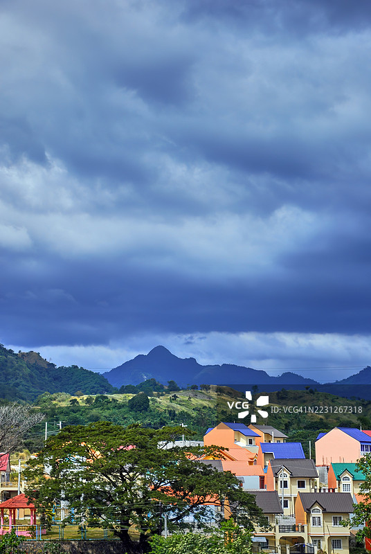 一片住宅区坐落在山丘和山脉的脚下，五彩斑斓的房屋在戏剧性的多云天空下显得格外迷人。图片素材