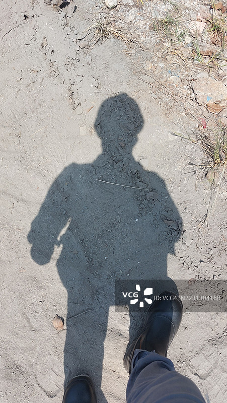 在明亮的正午阳光下，穿着靴子的人的完整影子投射在尘土飞扬的地面上。图片素材