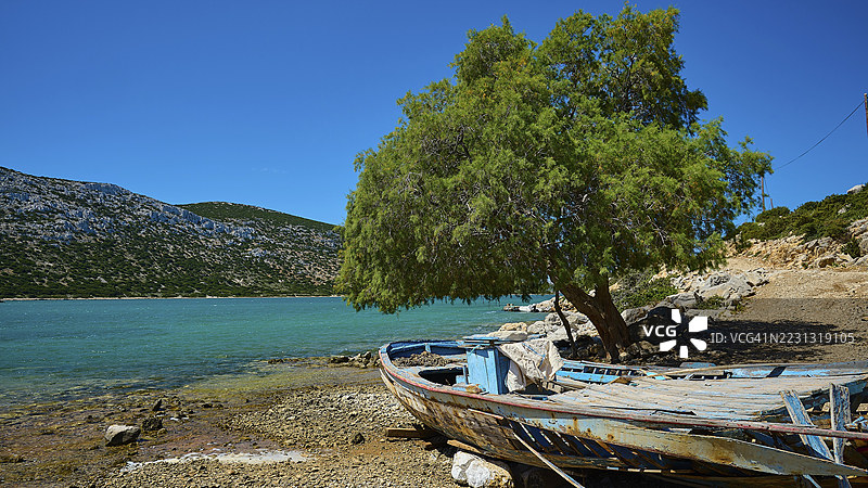 一艘破旧的船只停靠在宁静的海岸边,旁边有一棵大树,位于希腊多德卡尼斯群岛的瓦伊湾,阿斯提帕利亚。图片素材